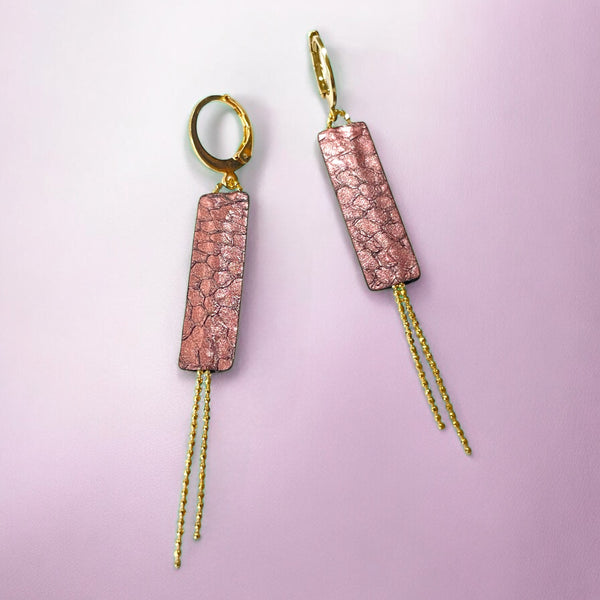 Boucles d'oreilles Effinis de Nune, dorées à l'or fin avec cuir marin, cuir de poisson saumon veux rose métallisé, de forme effilée avec chaînettes dorées pendantes, sur fond rose