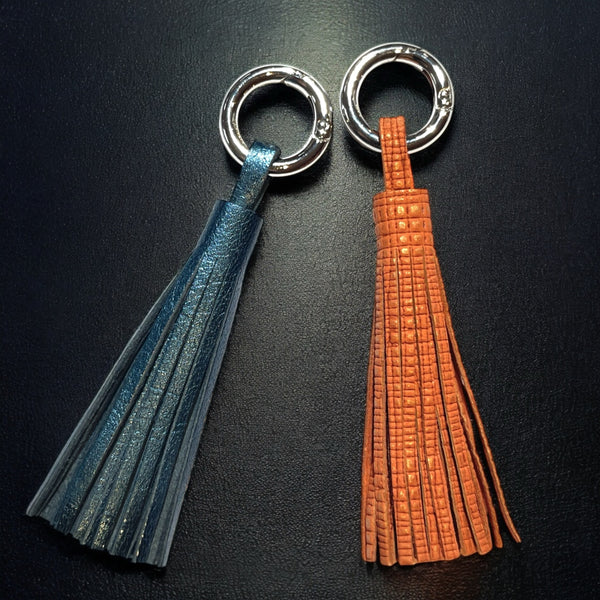 2 Bijoux de sac argentés, chacun portant un grand pompon en cuirs bleu nuit métallisé et orangé or mat, sur fond noir