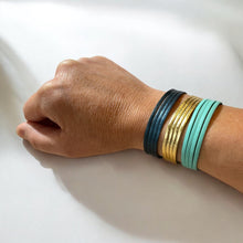 Charger l&#39;image dans la galerie, 3 bracelets à 4 liens en cuirs doré, bleu nuit métallisé et vert menthe, portés au poignet sur fond blanc
