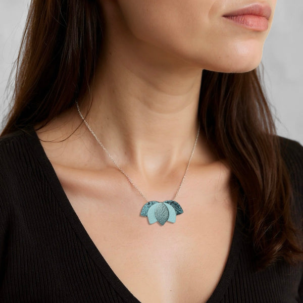 Collier argenté court et délicat avec un pendentif en forme de Lotus, composé de cuirs bleu paon pailleté, bleu nuage et bleu gris nacré, porté au cou d'un femme brune