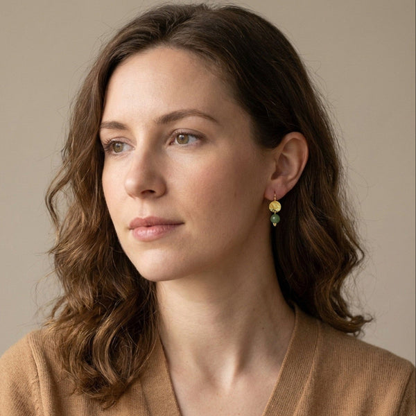 Petites boucles d'oreilles rondes en métal martelé doré à l'or fin avec pendant en perle d'opale verte, portées aux oreilles par une femme brune