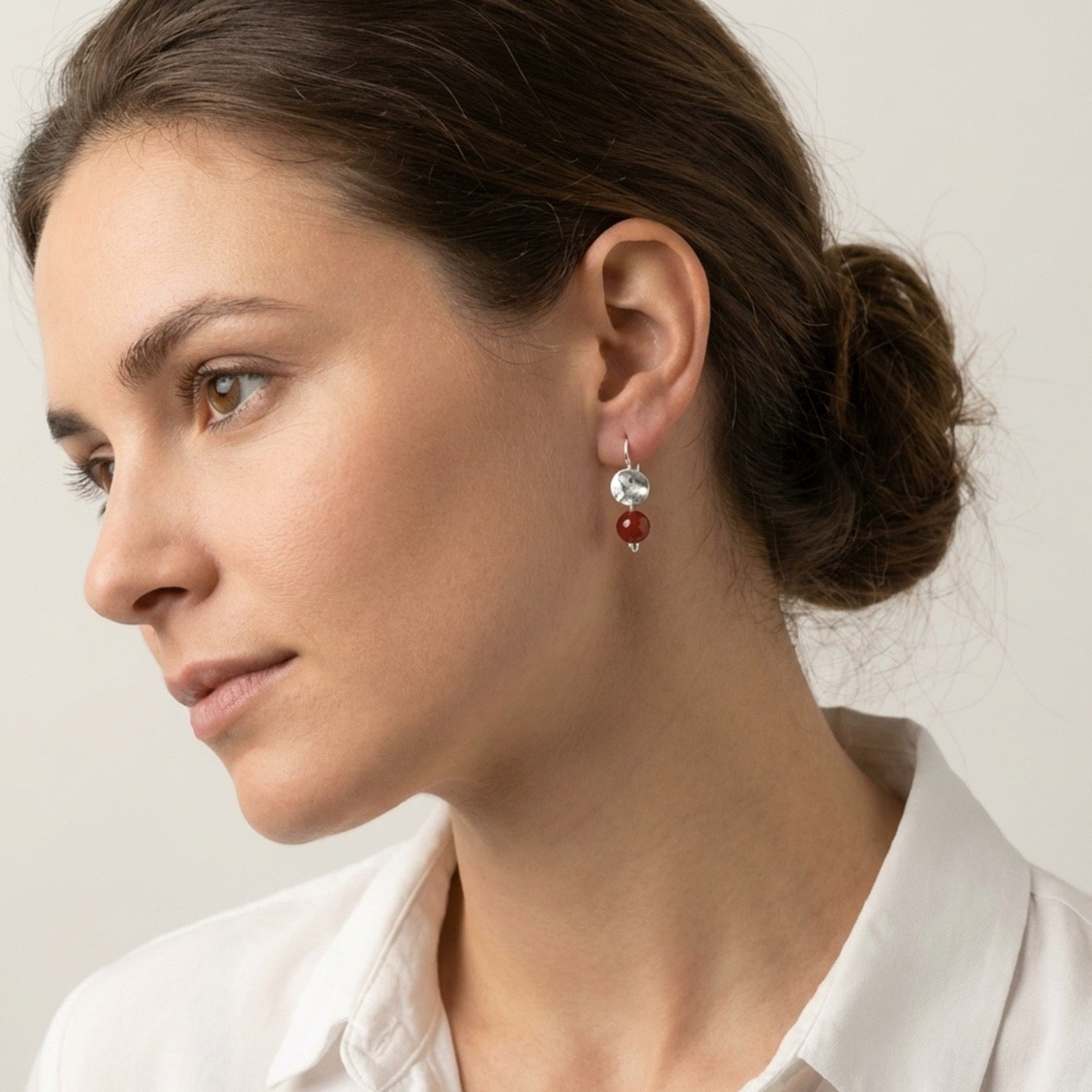 Petites boucles d'oreilles rondes en métal martelé doré à l'or fin avec pendant en perle de mokaïte bordeaux, portées à l'oreille par une femme brune
