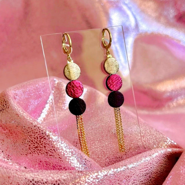 Longues et fines boucles d'oreilles Nune à franges dorées avec 3 petits disques de cuir recyclé cannelle dorée satinée, fuchsia métallisé et violine satinée sur fond cuir rose brillant. Modèle Janis