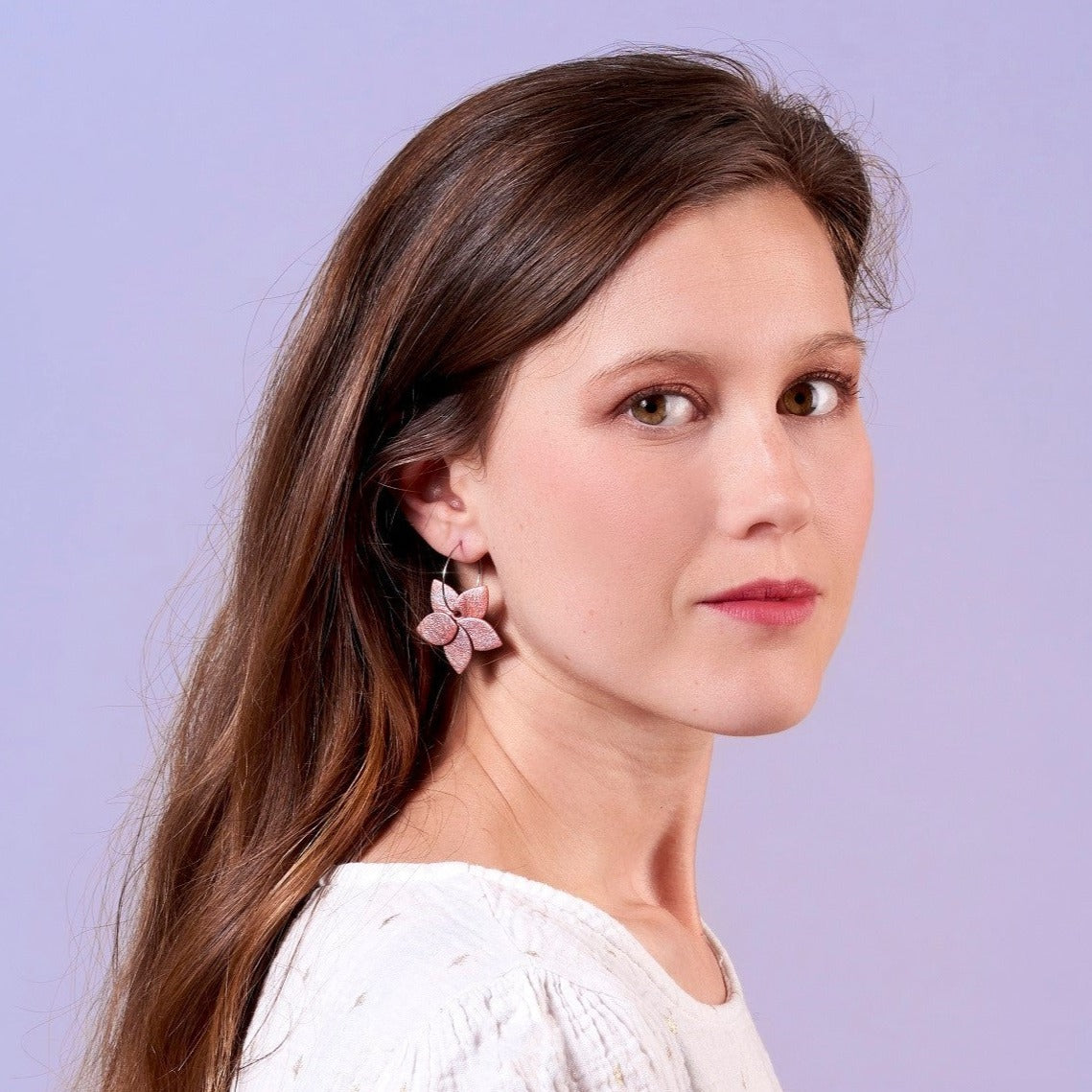 Jeune femme aux longs cheveux bruns portant les petites créoles Jasmin Argenté de Nune, boucles d'oreilles créoles en forme de fleurs en cuir rose nacré et anneaux en acier inoxydable argenté, sur fond mauve