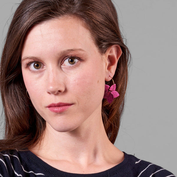 Jeune femme vue de face et de près portant les petites créoles Jasmin en pétales de cuir fushia métallisé, en forme de fleurs, sur fond gris