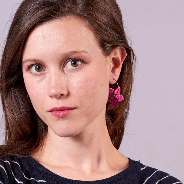 Jeune femme vue de face et de près portant les petites créoles Jasmin en pétales de cuir fushia métallisé, en forme de fleurs, sur fond mauve