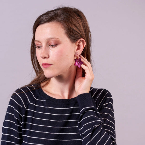 Jeune femme vue de 3/4 portant les petites créoles Jasmin en pétales de cuir fushia métallisé, en forme de fleurs, sur fond mauve