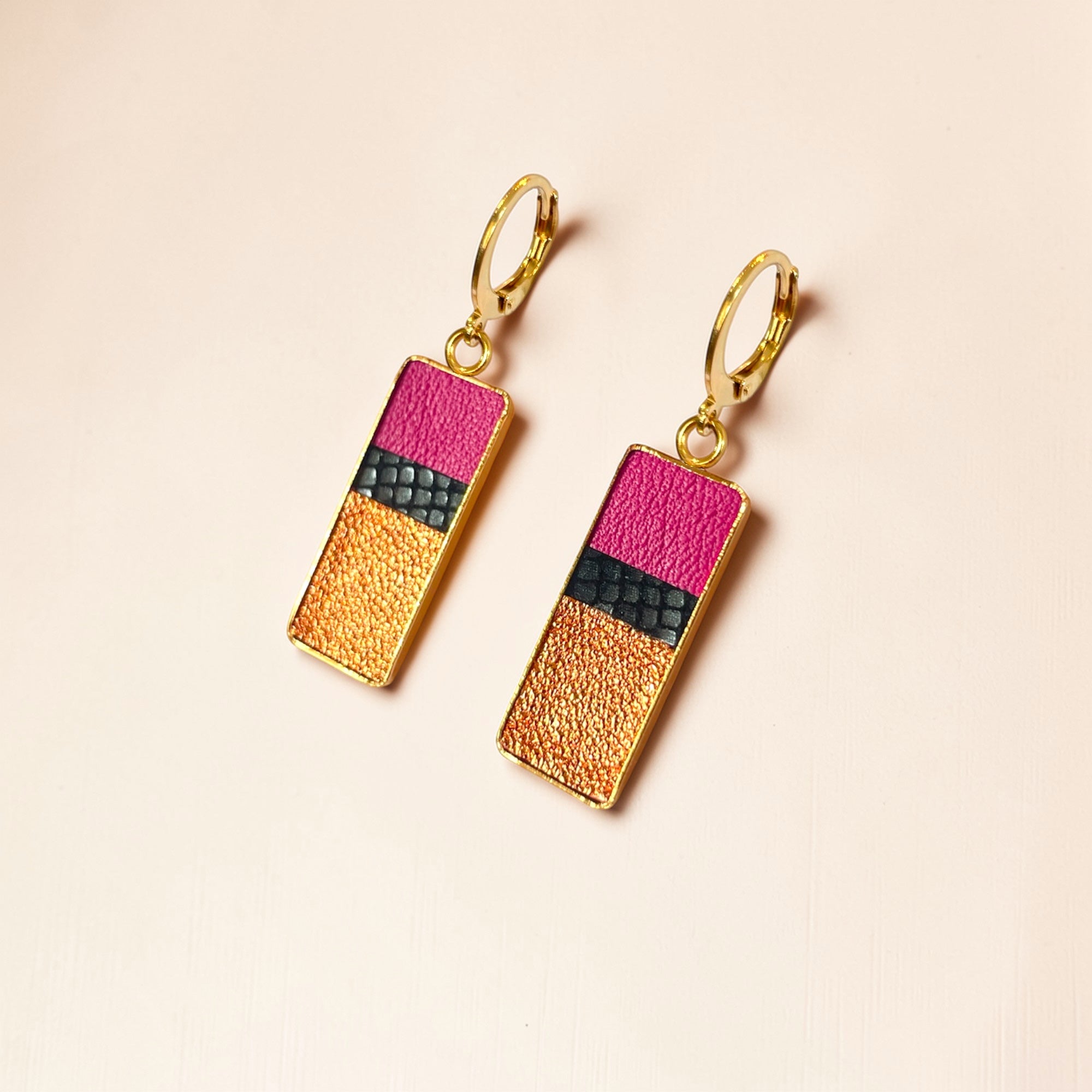 Petites boucles d'oreilles dorées pendantes rectangulaires verticales tricolores en cuirs rose fushia, noir petit grain irisé et cuivre clair, posées à plat sur fond orangé
