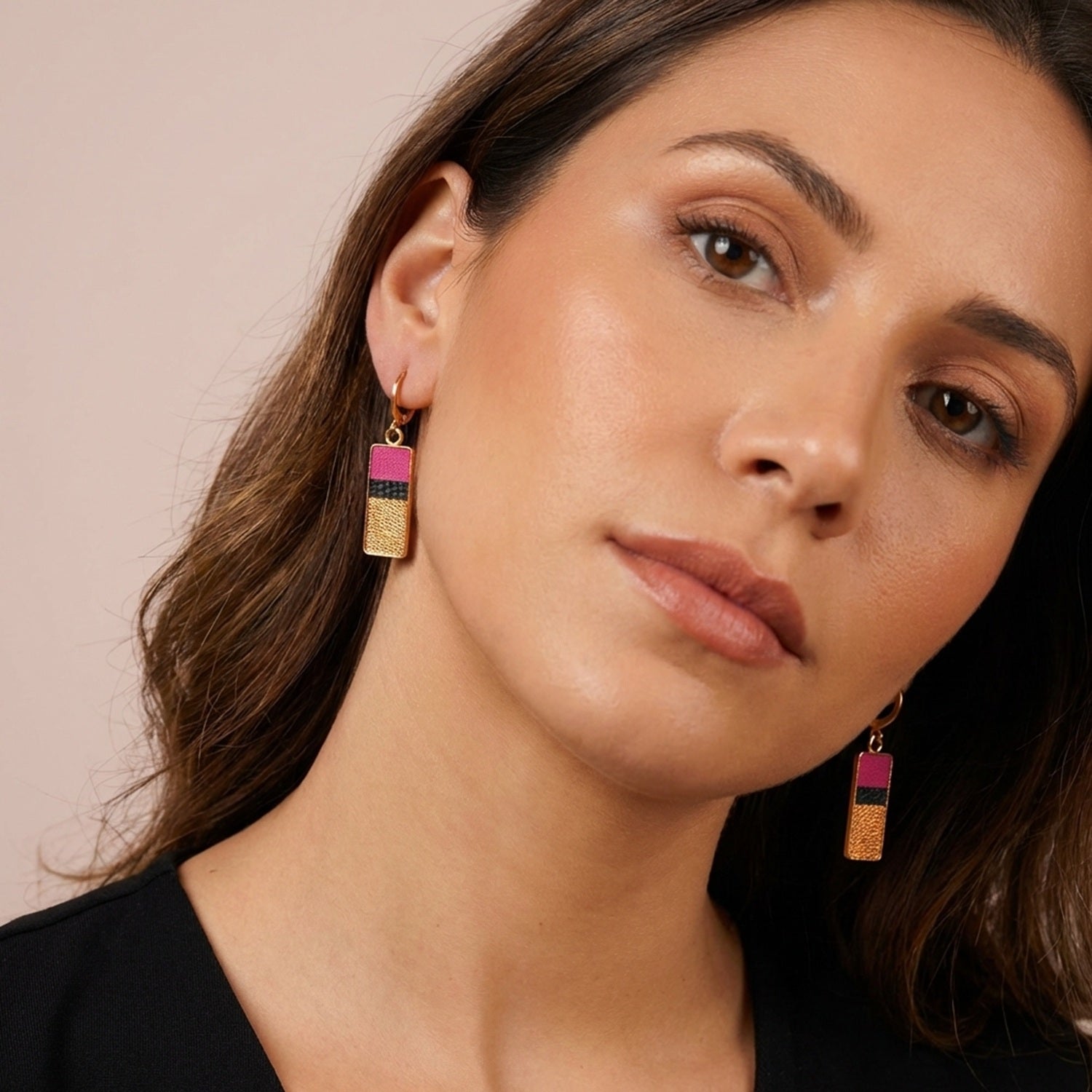 Petites boucles d'oreilles dorées pendantes rectangulaires verticales tricolores en cuirs rose fushia, noir petit grain irisé et cuivre clair, portées au oreilles par une femme brune aux cheveux lâchés