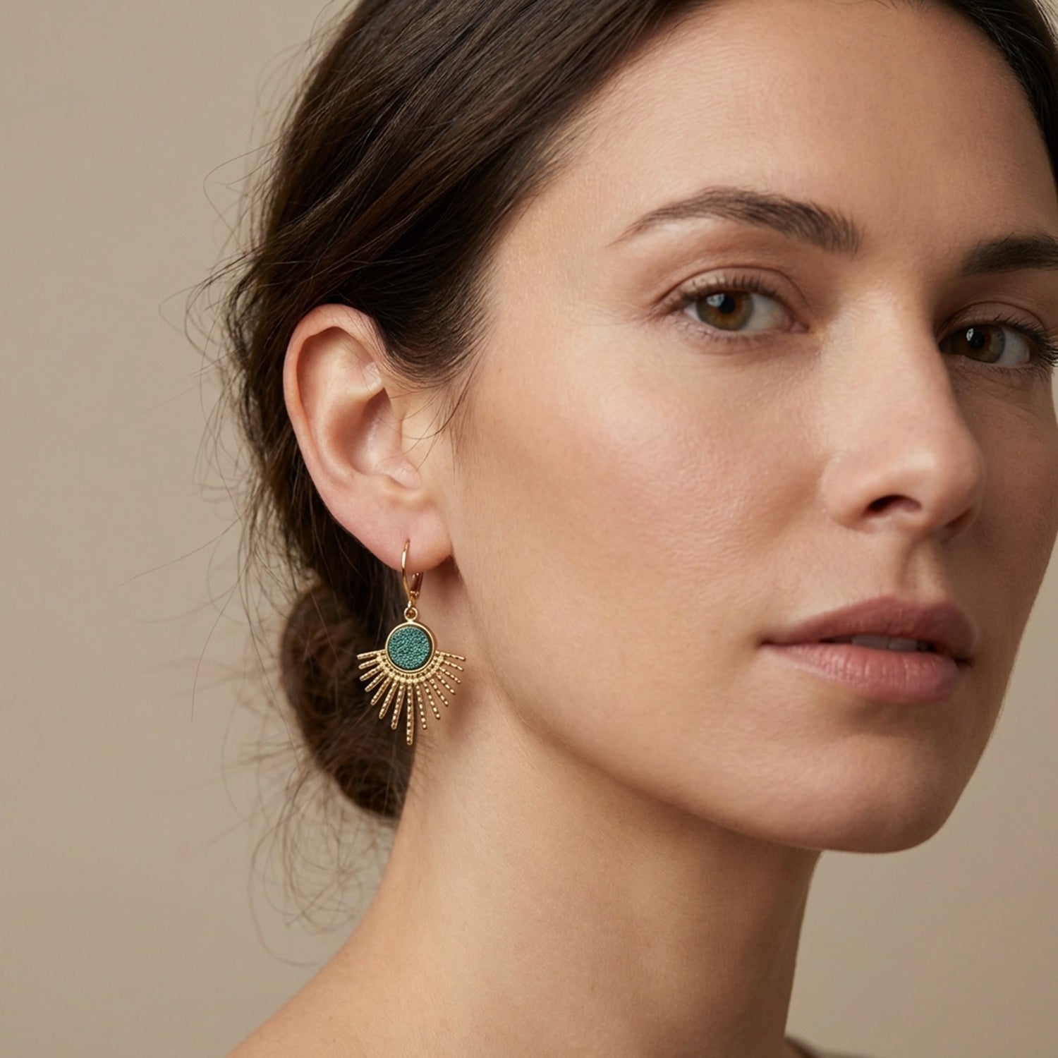 Petites boucles d'oreilles dorées Miria forme soleils avec cabochons de cuir bleu paon pailleté, portées au oreilles par une femme brune
