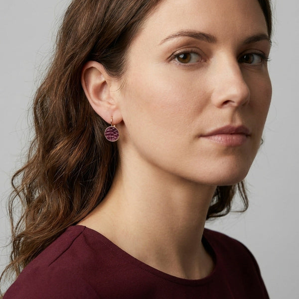 Petites boucles d'oreilles dormeuses dorées serties de fin cuir marin grenat métallisé, portées à l'oreille par une femme brune