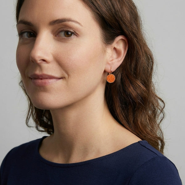 Petites boucles d'oreilles dormeuses dorées serties de fin cuir marin orange métallisé, portées à l'oreille par une femme brune