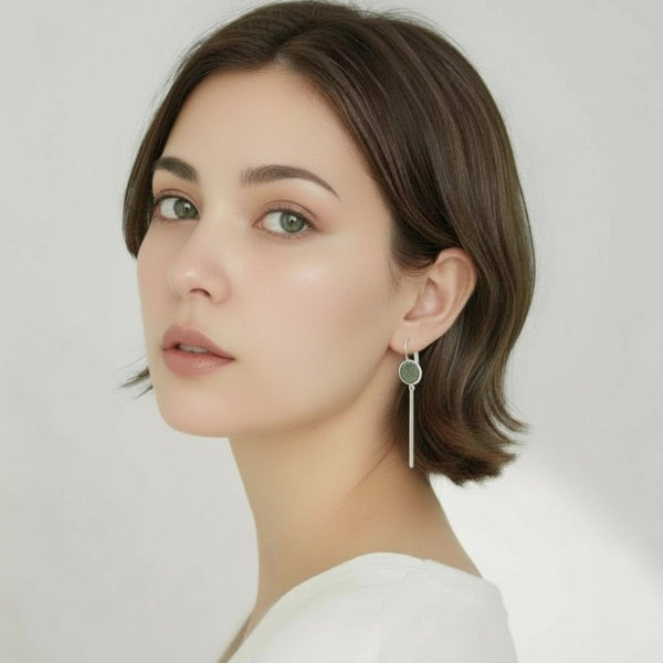 Jeune femme vue de près portant les Longues et fines boucles d'oreilles argentées Naka avec cabochon en cuir kaki métallisé, sur fond gris