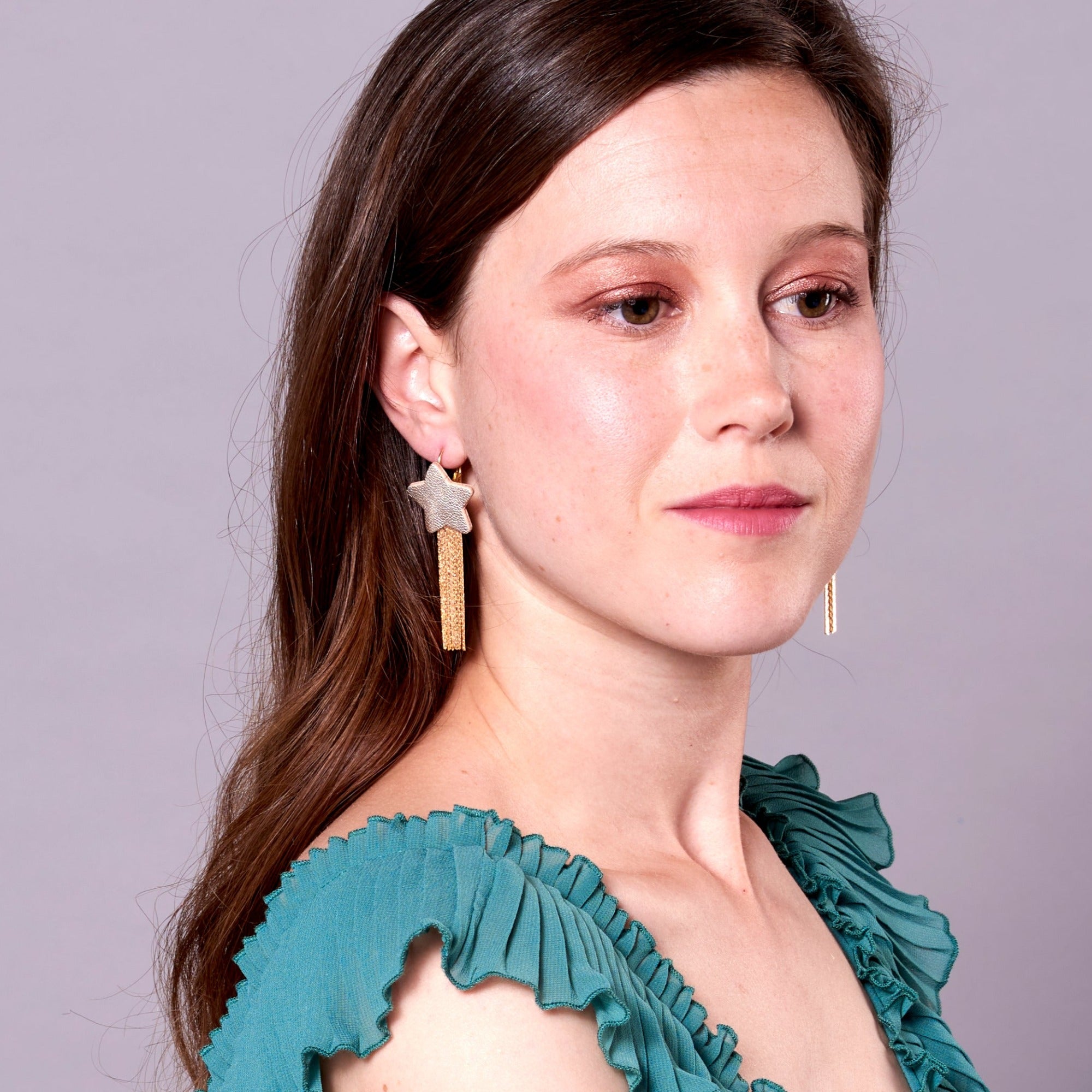Jeune femme portant les Boucles d'oreilles légères SuperStella, grandes étoiles en cuir argenté recyclé et leurs longues et fines chaînettes dorées, sur fond mauve