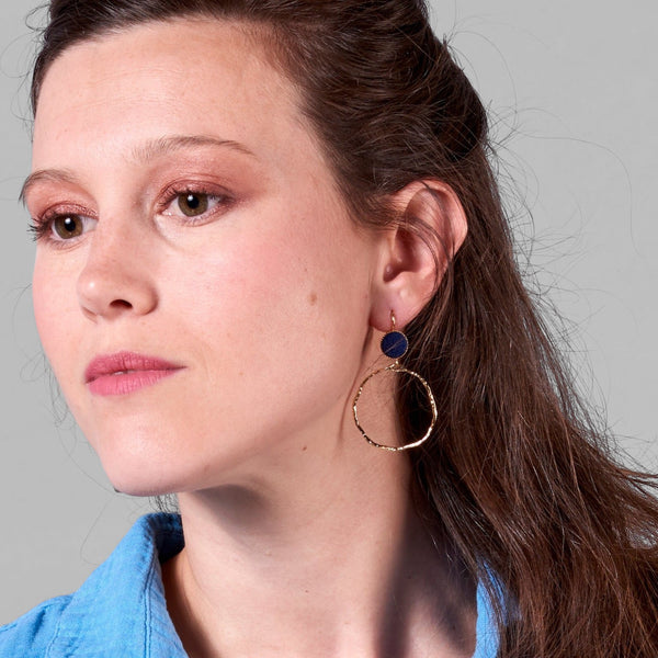 Jeune femme vue de 3/4 portant les boucles d'oreilles dorées Nune modèle Bali Créoles en cuir marine satiné, sur fond gris