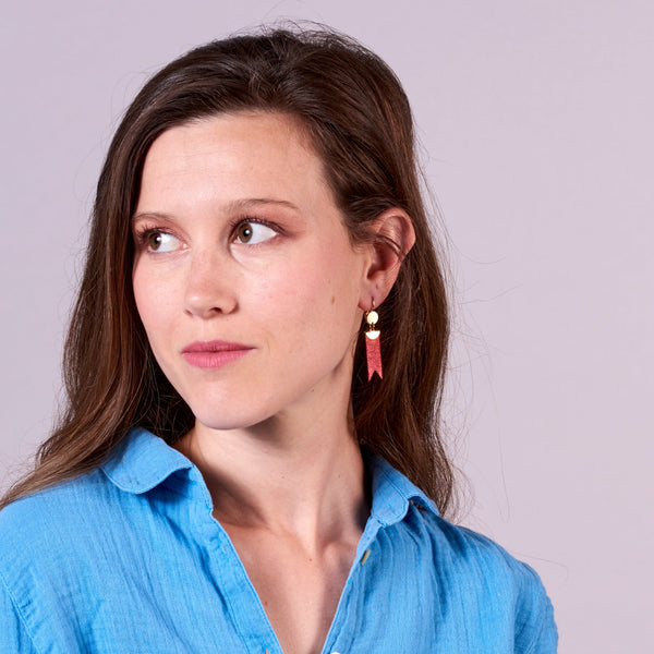 Jeune femme portant les Boucles d'oreilles dorées Nune modèle Cinta en forme de rubans en cuir rubis métallisé, sur fond gris