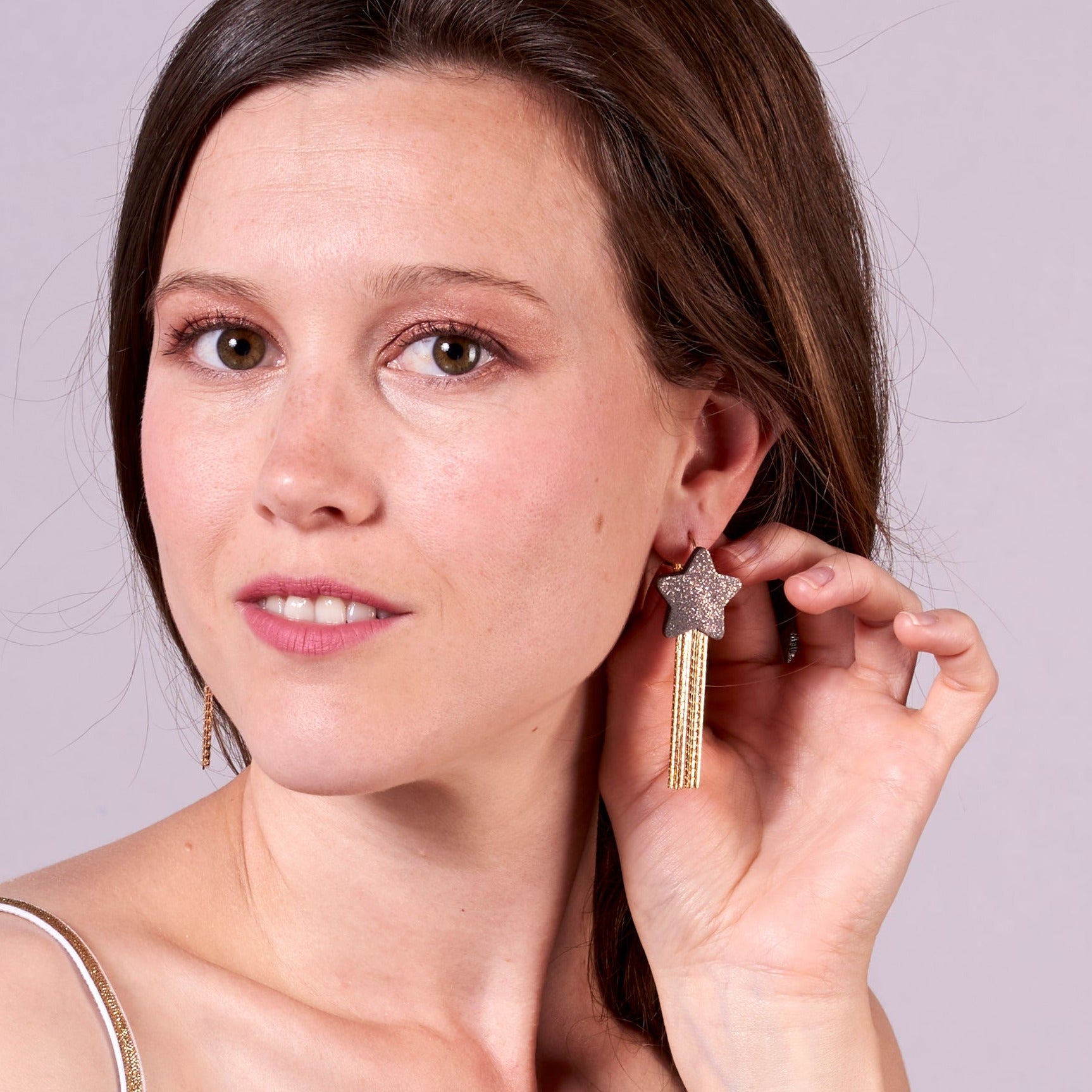 Jeune femme vue de tout près portant les Boucles d'oreilles légères SuperStella cuir pailleté, grandes étoiles en cuir gris pailleté irisé recyclé et leurs longues et fines chaînettes dorées, sur fond mauve