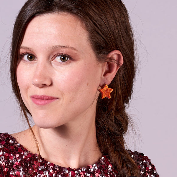 Jeune fille aux longs cheveux bruns vue de 3/4 portant les boucles d'oreilles Diva de Nune en forme d'étoile en cuir recyclé orange sanguine métallisé, sur mauve