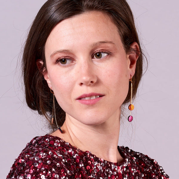 Jeune fille vue de près portant les Longues et fines boucles d'oreilles Nune dorées terminées petits disques de cuir recyclé orange sanguine métallisé et fushia métallisé. Modèle Merry
