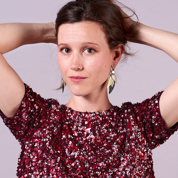 Jeune femme tenant ses cheveux bruns portant les boucle d'oreille dorée Nune modèle Ara en plumes de cuir argent, doré brillant et noir grainé irisé, sur fond mauve
