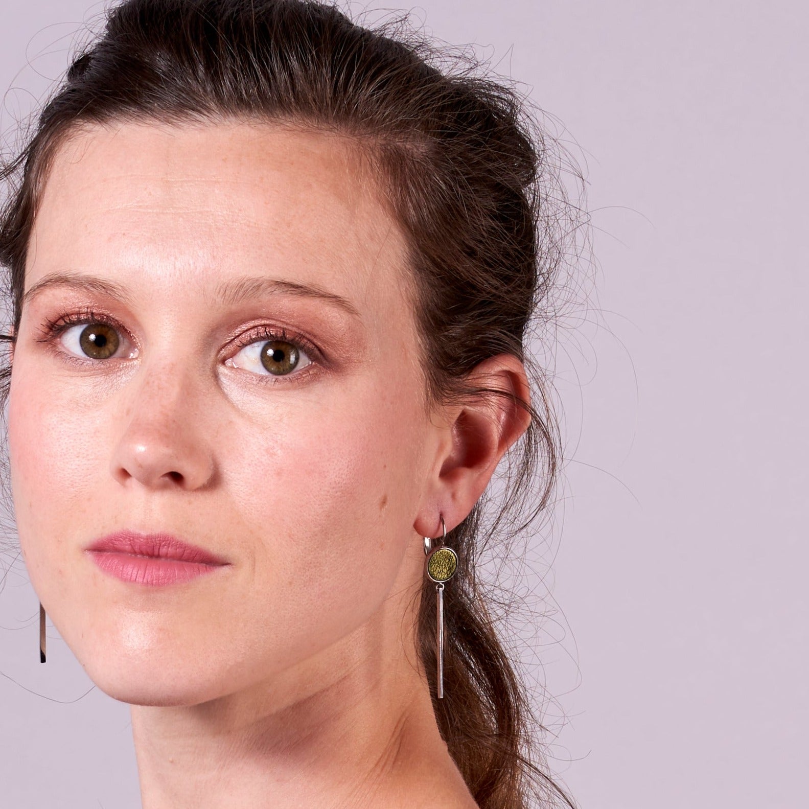 Jeune femme vue de près portant les Longues et fines boucles d'oreilles argentées Naka avec cabochon en cuir kaki métallisé, sur fond gris