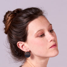 Charger l&#39;image dans la galerie, Jeune femme à chignon vue de profil portant les boucles d&#39;oreilles rondes bicolores Lumia en cuir noir pailleté irisé et noir grainé, sur fond mauve
