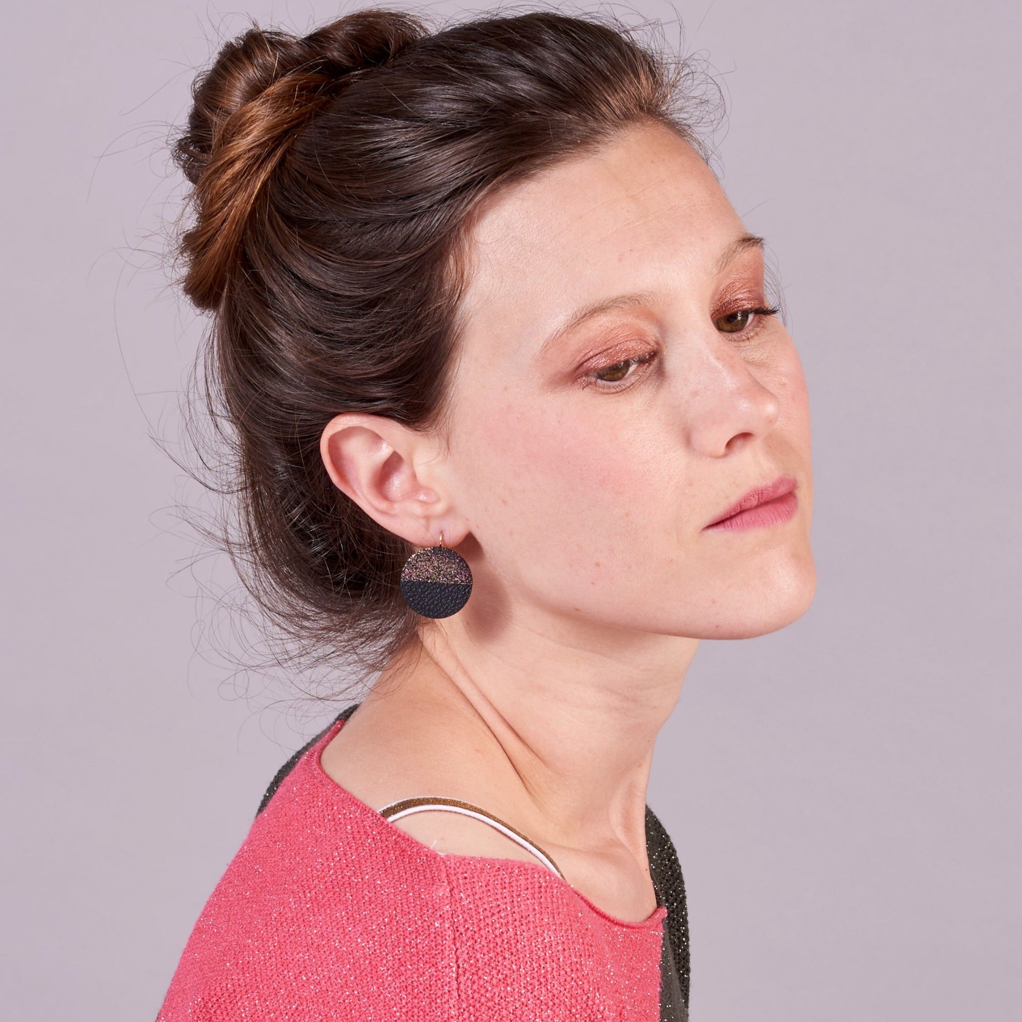 Jeune femme à chignon vue de 3/4 portant les boucles d'oreilles rondes bicolores Lumia en cuir noir pailleté irisé et noir grainé, sur fond mauve