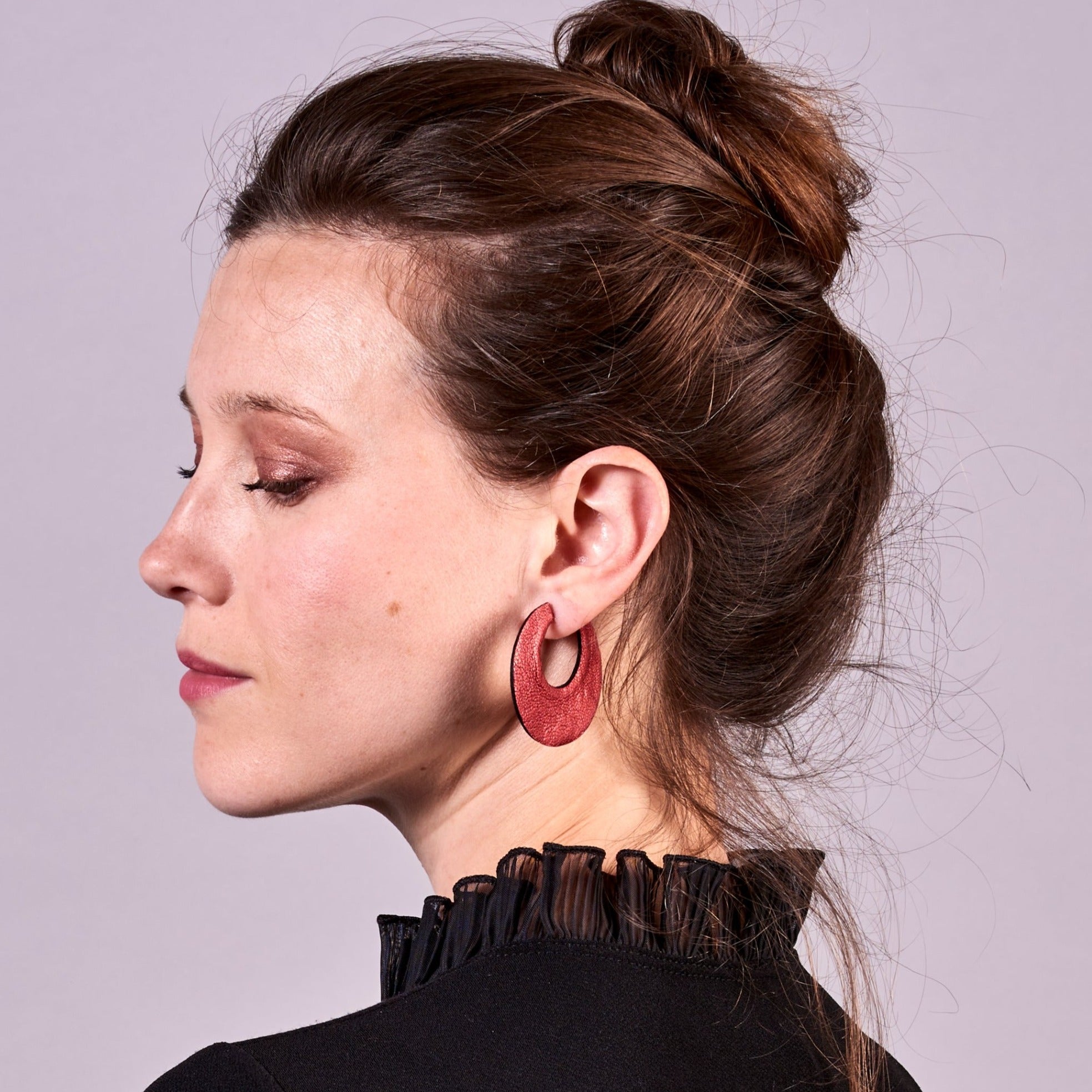Jeune femme à chignon vue de profil portant les grandes boucles d'oreilles créoles rondes tout-cuir Circus, vues du côté rubis métallisé