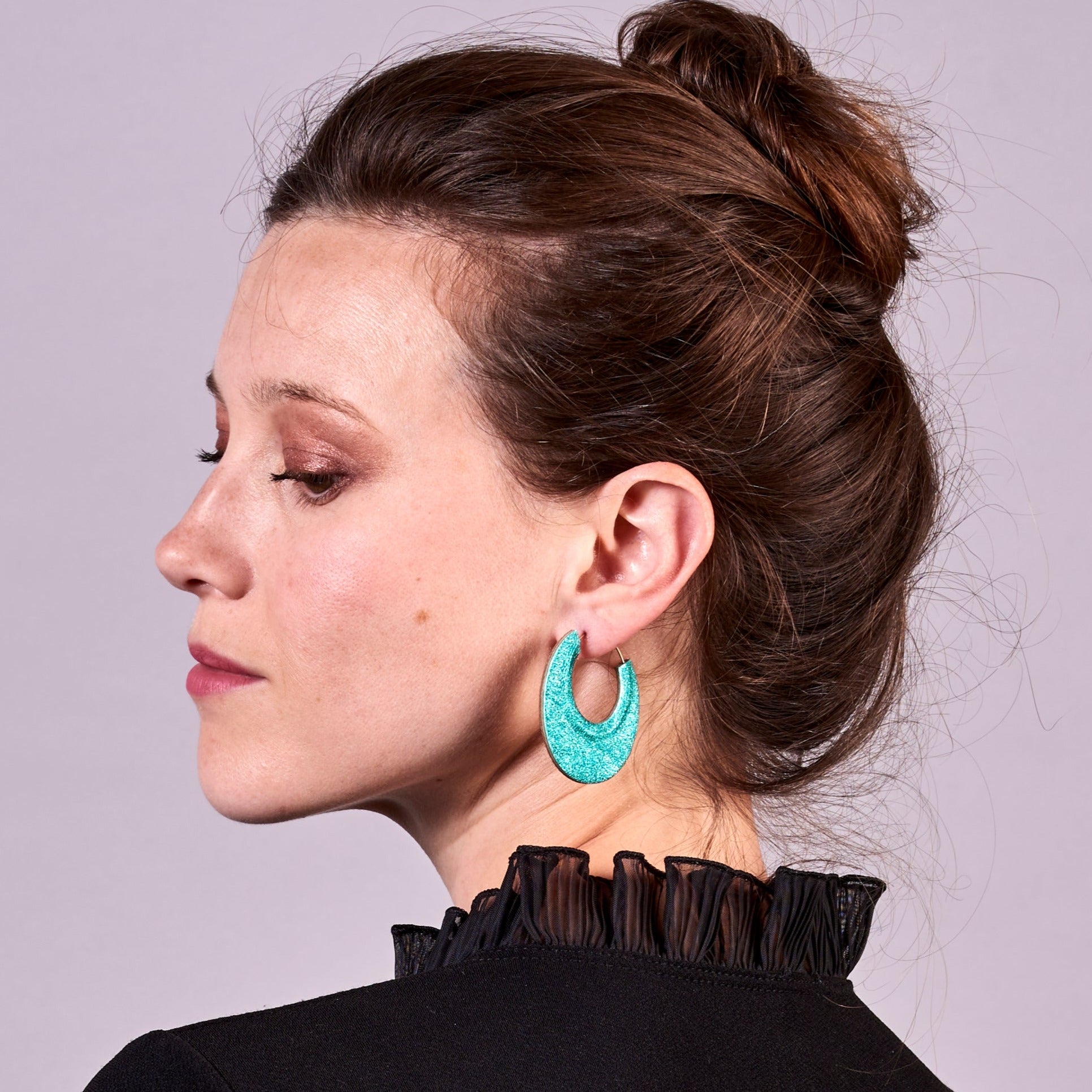 Jeune femme à chignon vue de profil portant les grandes boucles d'oreilles créoles rondes tout-cuir Circus, vues du côté turquoise métallisé