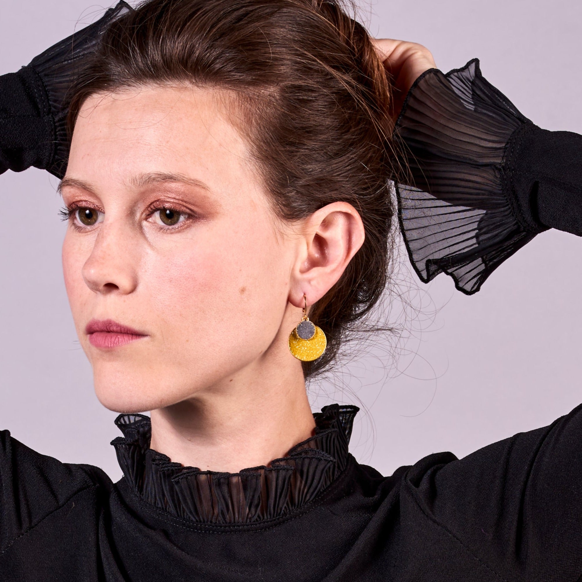 Jeune femme brune vue de près relevant ses cheveux portant les Petites boucles d'oreilles dorées rondes Bulles Pailletées de Nune avec doubles sertis de cuirs pailletés safran et gris, sur fond mauve