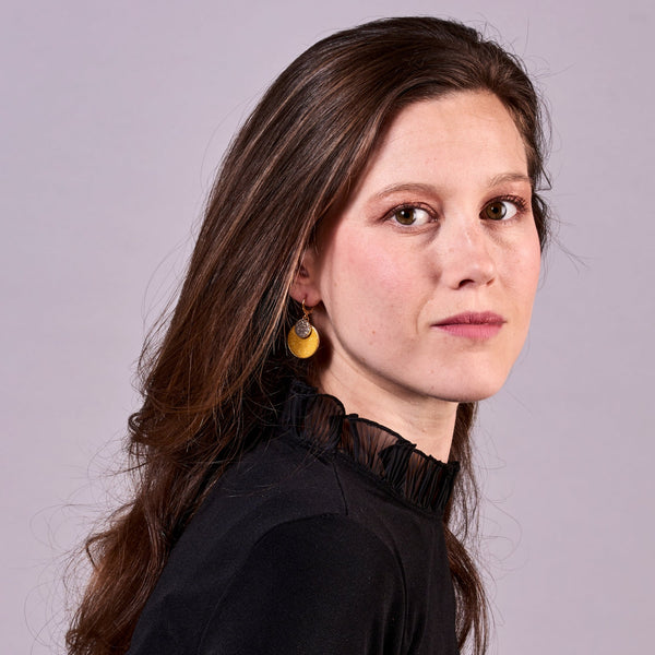 Jeune femme aux longs cheveux bruns vue de profil portant les Petites boucles d'oreilles dorées rondes Bulles Pailletées de Nune avec doubles sertis de cuirs pailletés safran et gris, sur fond mauve