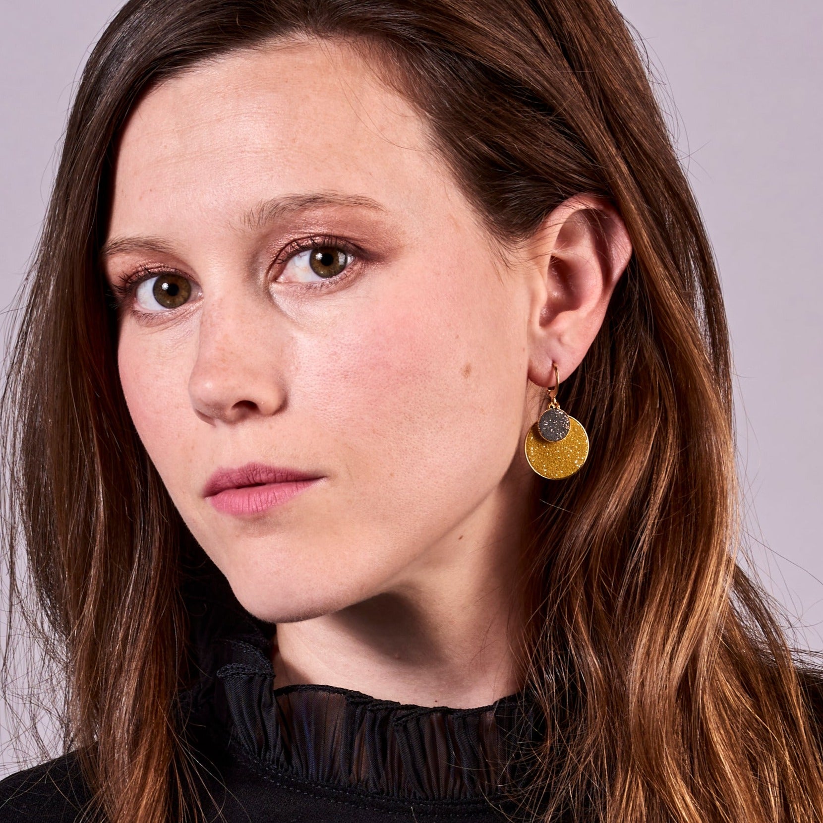 Jeune femme brune vue de près portant les Petites boucles d'oreilles dorées rondes Bulles Pailletées de Nune avec doubles sertis de cuirs pailletés safran et gris, sur fond mauve