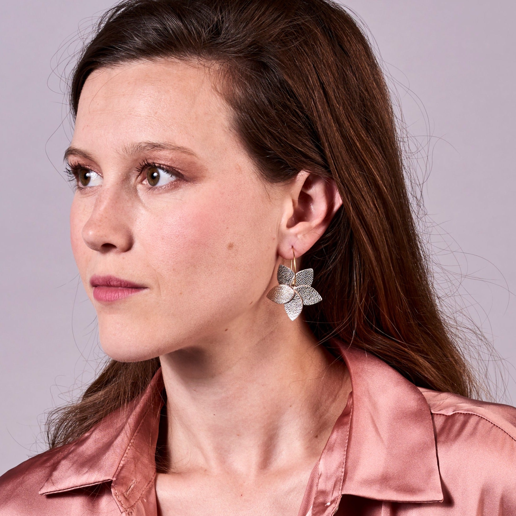 Jeune femme vue de profil portant les petites créoles Jasmin en pétales de cuir argent, en forme de fleurs, sur fond mauve