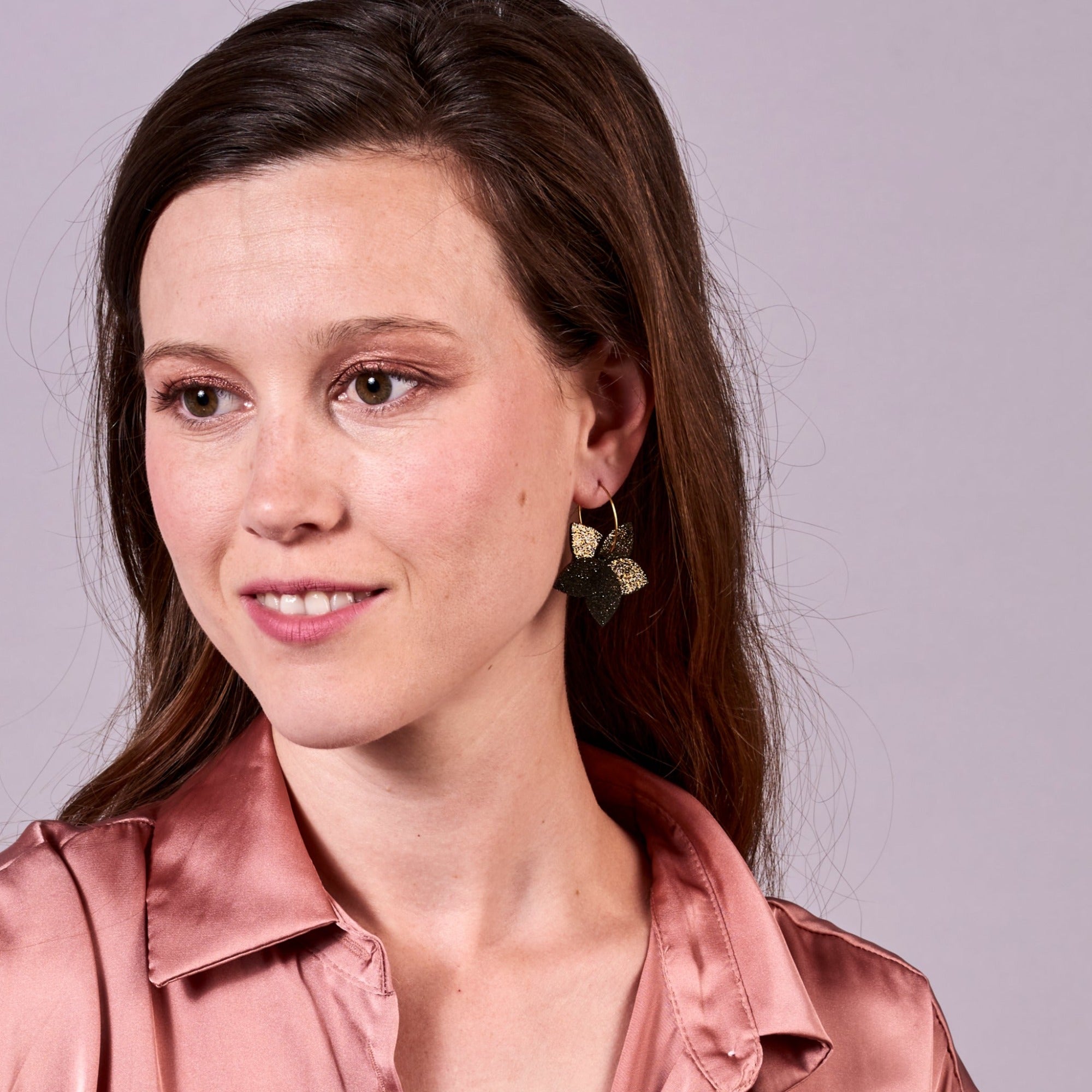 Jeune femme vue de 3/4 portant les petites créoles Jasmin en pétales de cuir noir pailleté or, en forme de fleurs, sur fond mauve