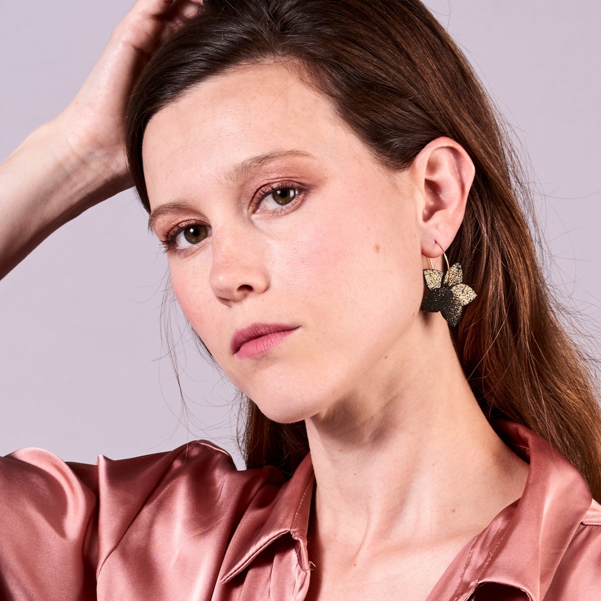 Jeune femme vue de face et de près portant les petites créoles Jasmin en pétales de cuir noir pailleté or, en forme de fleurs, sur fond mauve