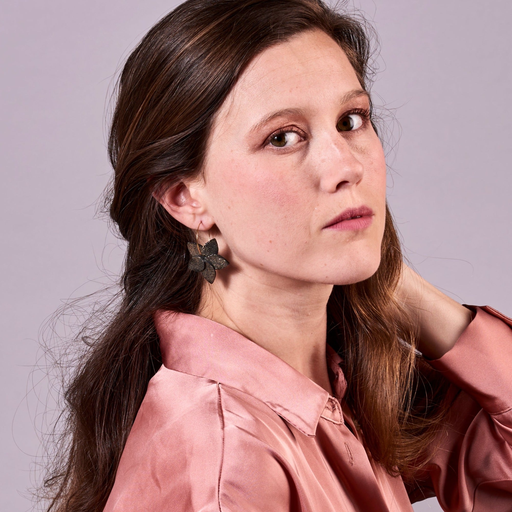 Jeune femme vue de profil portant les petites créoles Jasmin en pétales de cuir noir pailleté or, en forme de fleurs, sur fond mauve