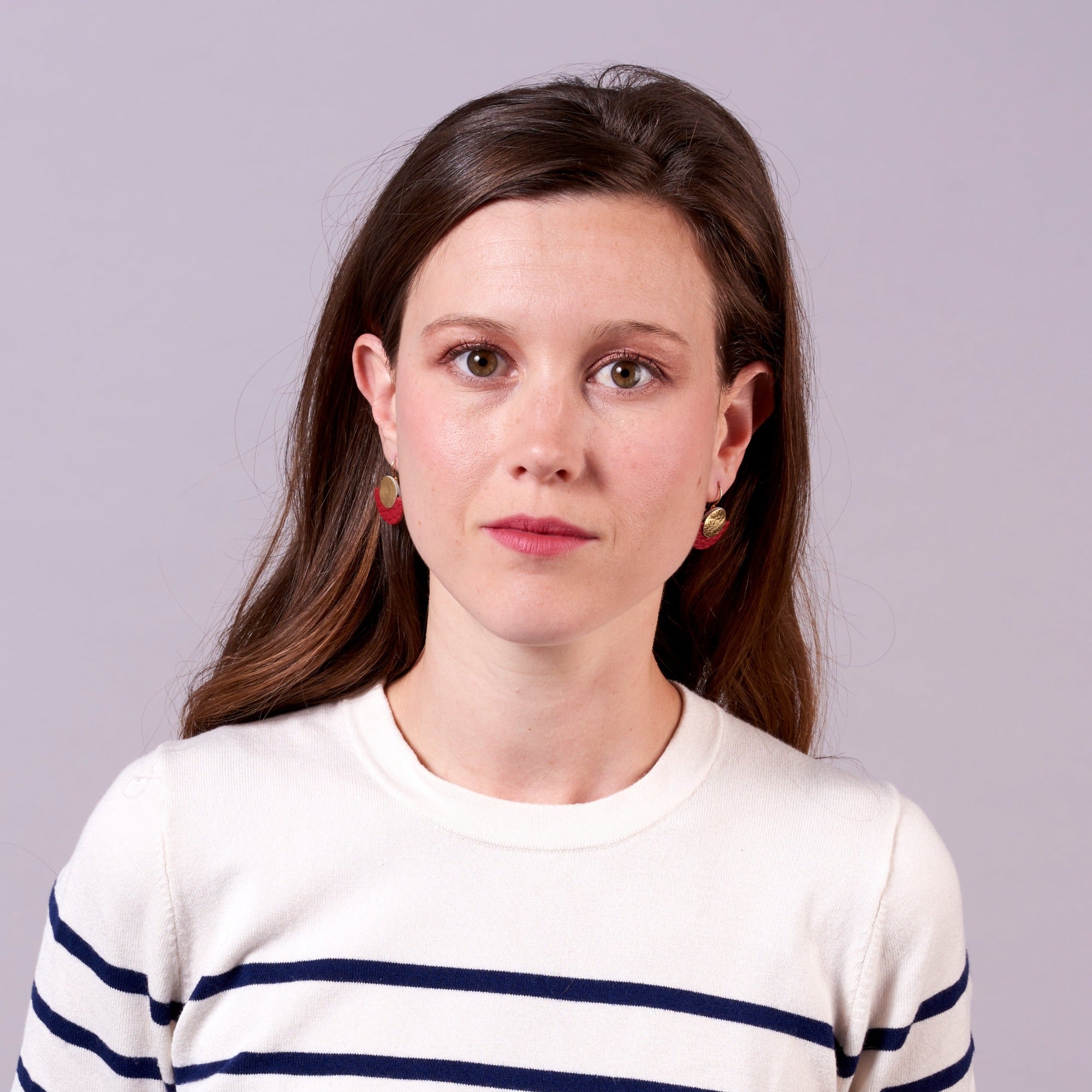 Jeune femme vue de buste portant les boucles d'oreilles Téthys dorées en forme de demi-lunes en cuirs doré et rouge grainé, sur fond mauve