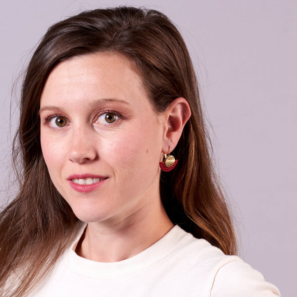 Jeune femme vue de près portant les boucles d'oreilles Téthys dorées en forme de demi-lunes en cuirs doré et rouge grainé, sur fond mauve