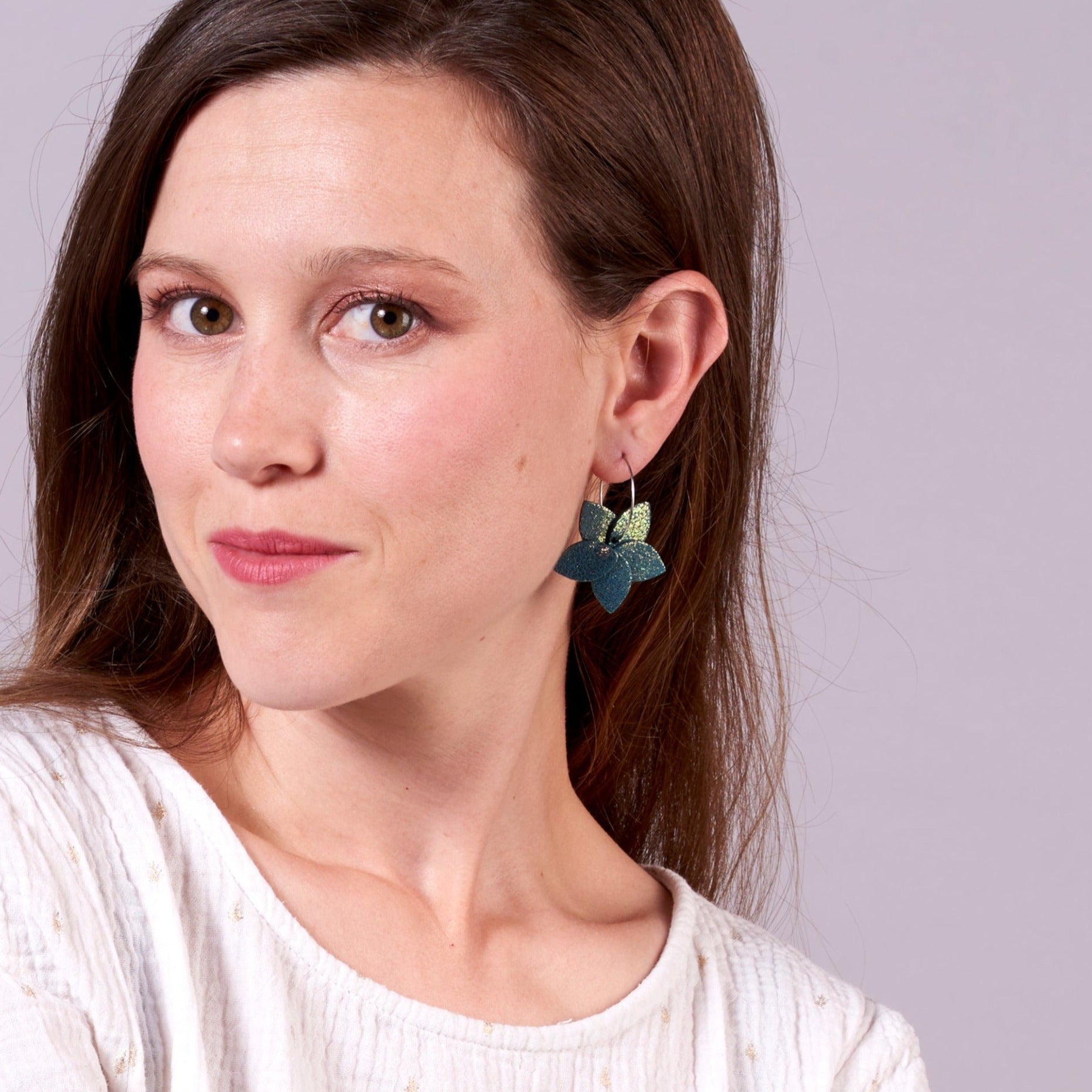 Jeune femme vue de 3/4 et de près portant les petites créoles Jasmin Argenté en pétales de cuir vert canard pailleté irisé, en forme de fleurs, sur fond mauve