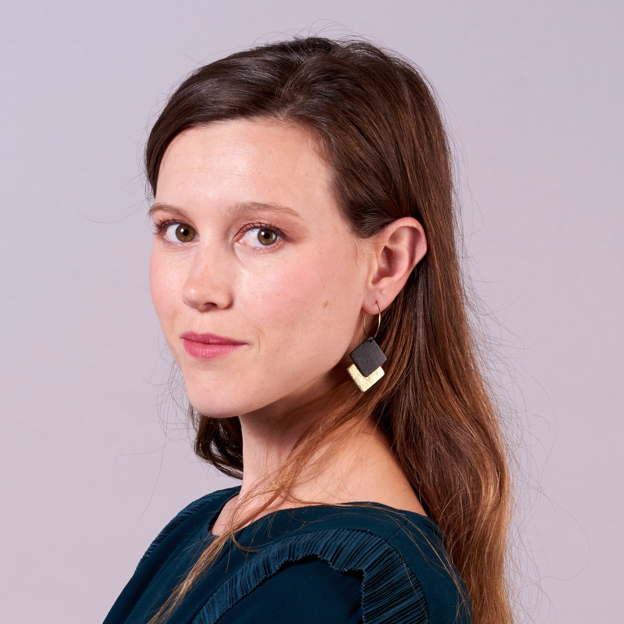 Jeune femme vue de 3/4 portant les boucles d'oreilles créoles Adalis dorées et duo de losanges en cuir recyclé noir satiné et doré sur fond gris rosé