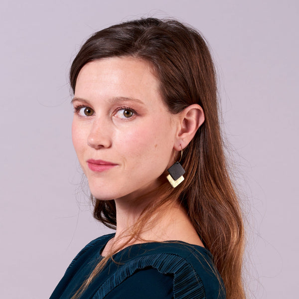 Jeune femme vue de 3/4 portant les boucles d'oreilles créoles Adalis dorées et duo de losanges en cuir recyclé noir satiné et doré sur fond gris rosé