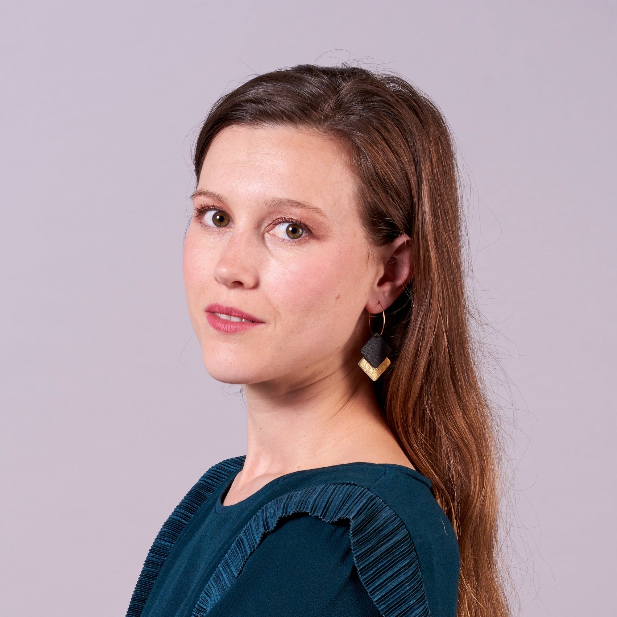 Jeune femme vue de 3/4 portant les boucles d'oreilles créoles Adalis dorées et duo de losanges en cuir recyclé noir satiné et doré sur fond mauve