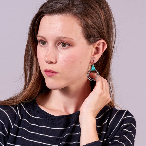 Jeune femme portant les Créoles argentée du modèle Vega de la marque Nune, avec une touche de cuir turquoise métallisé, sur fond mauve