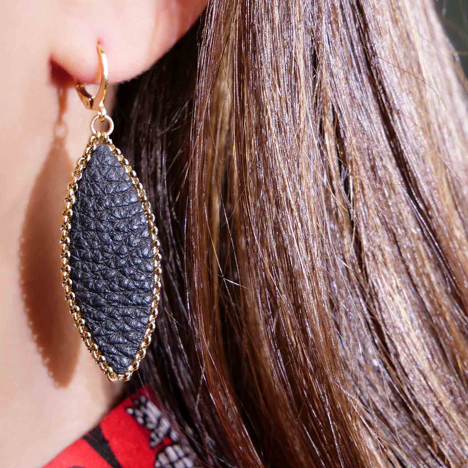 Boucle d'oreille pendante Nune modèle Auris en forme d'épi en cuir noir grainé cerclé de chaîne dorée, portée à l'oreille, vue en gros plan