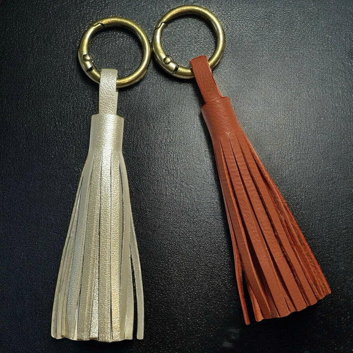 2 Bijoux de sac bronze, chacun portant un grand pompon en cuirs doré clair nacré et terracotta, sur fond noir