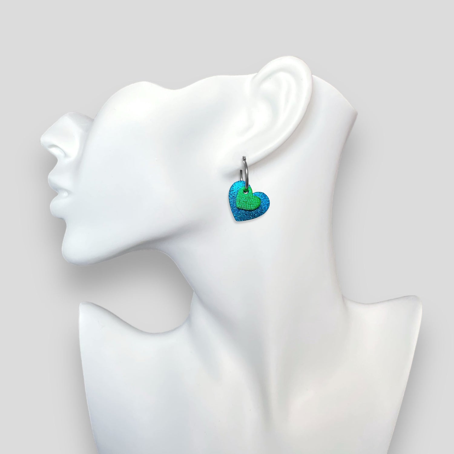 Boucles créoles argentées avec pendentifs en forme de cœurs bleu roi métallisé et vert métallisé, portées à l'oreille sur un buste blanc