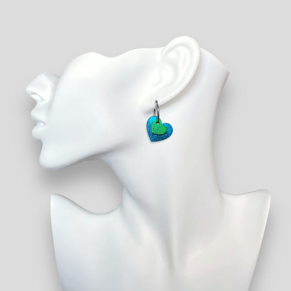 Boucles créoles argentées avec pendentifs en forme de cœurs bleu roi métallisé et vert métallisé, portées à l'oreille sur un buste blanc