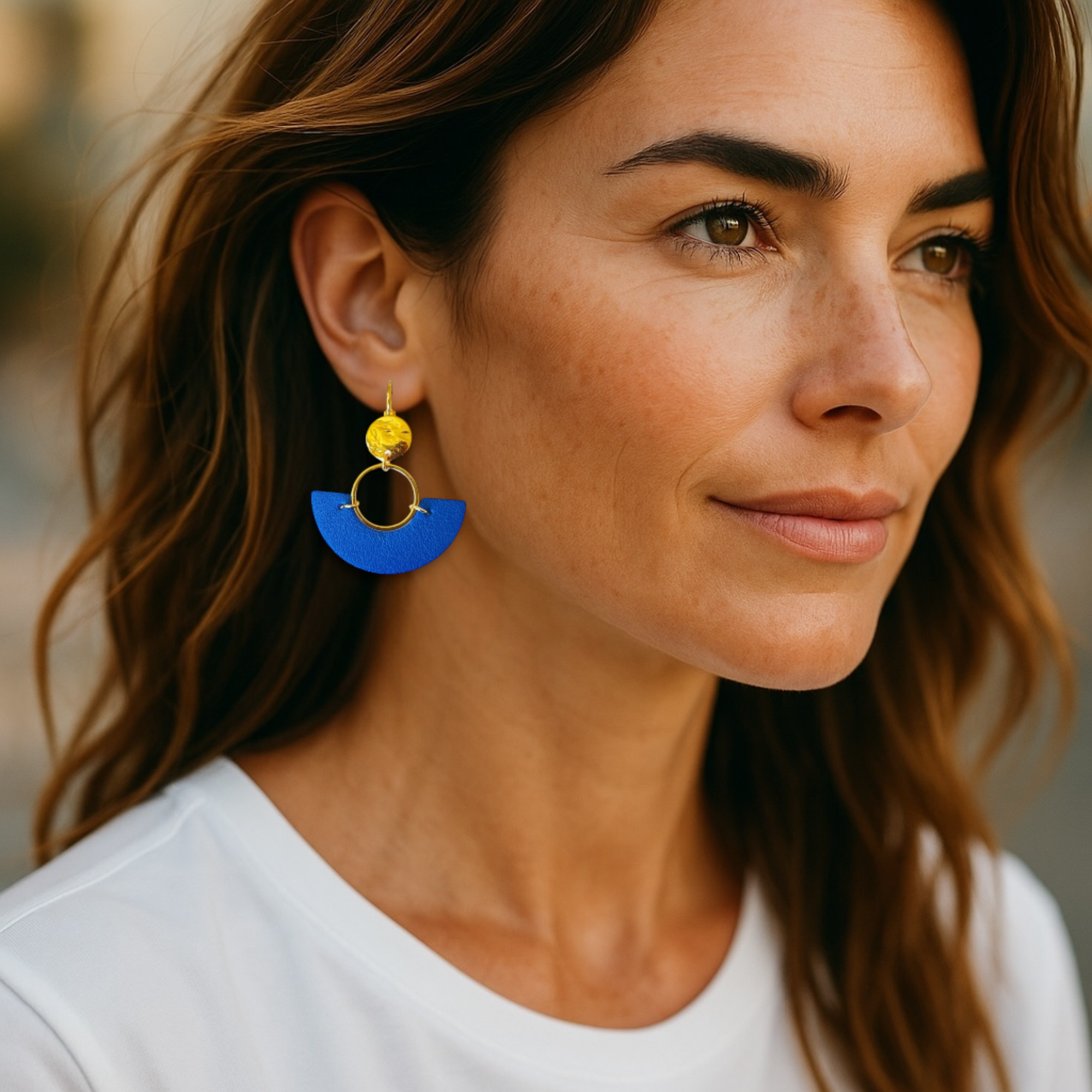 boucles d'oreilles dorées pendantes en forme de demi-lunes en cuir bleu roi, portées à l'oreille par une femme brune