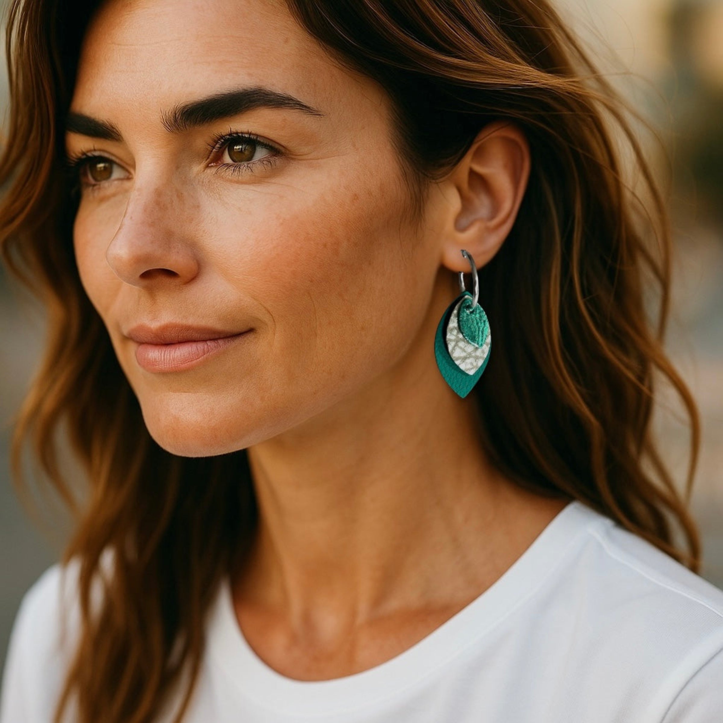 Créoles argentées avec pendentifs modulables en forme de feuilles en cuirs vert émeraude métallisé, argent grainé et vert émeraude grainé, portées à l'oreille par une femme brune