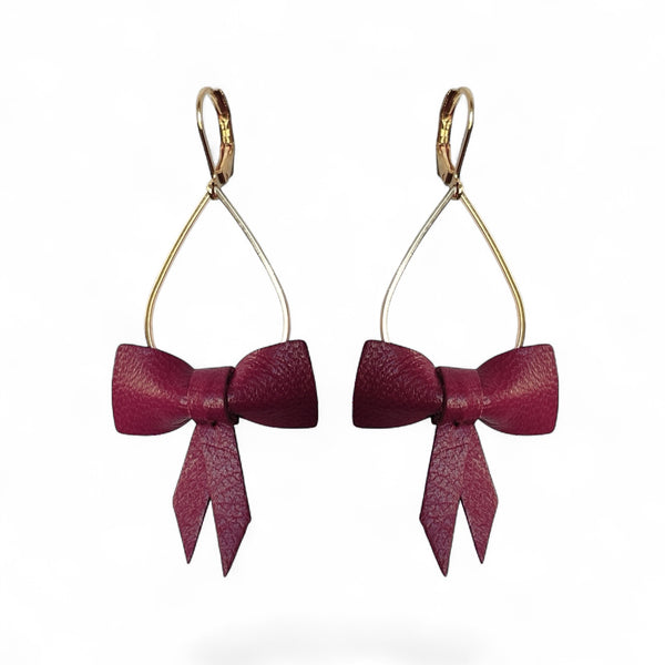Boucles d'oreilles dorées pendantes et légères en forme de gouttes, décorées de délicats nœuds de rubans de cuir pourpre, sur fond blanc. Modèle Séraphine de Nune.